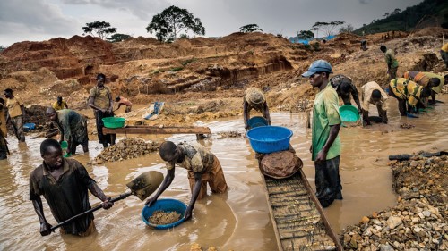 A group of coltan miners in the Congo stand waist deep in brown, muddy water as they use shovels to remove coltan from the water.