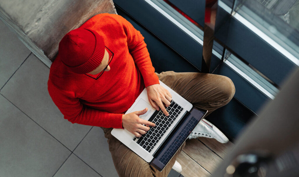 Above view of young freelancer man in glasses wear red hat and wool sweater sitting on bench next by window legs crossed, remote online working on laptop, typing on keyboard. Distance job.
